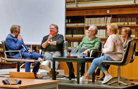 Eine fünfköpfige Gruppe sitzt nebeneinander auf einer kleinen Bühne im Lesesaal der Wissenschaftlichen Bibliothek und spricht miteinander. Im Hintergrund sind zahlreiche Bücher zu sehen.