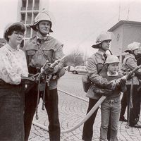 Ein Mädchen und ein Junge halten gemeinsam mit Feuerwehrmännern Feuerwehrschläuche.