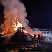 Das Bild zeigt einen brennenden Holzstapel mitten in der Nacht. Es ist dunkel und man sieht helle Flammen. Feuerwehrleute löschen mit Schläuchen, die auf den Brand gerichtet sind.