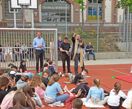 Viele Jugendliche sitzen auf dem Sportplatz-Boden während der Pater den Platz mit Weihwasser einsegnet