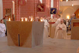 Mehrere Geistliche in weißen Gewändern stehen und knien vor einem Altar. Im Hintergrund sind weitere Kirchenbesucher zu sehen.