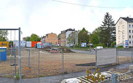 Abgezäunte Straßenbaustelle mit großer geschotterter Fläche, im Hintergrund sind Mehrfamilienhäuser zu sehen.